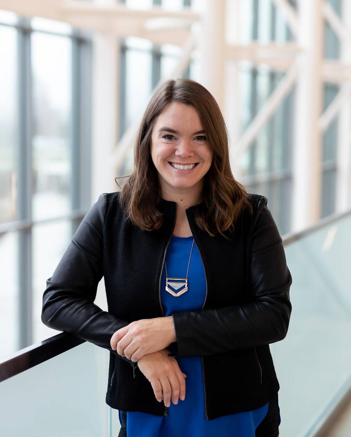 female in blue shirt and black coat standing in library smiling